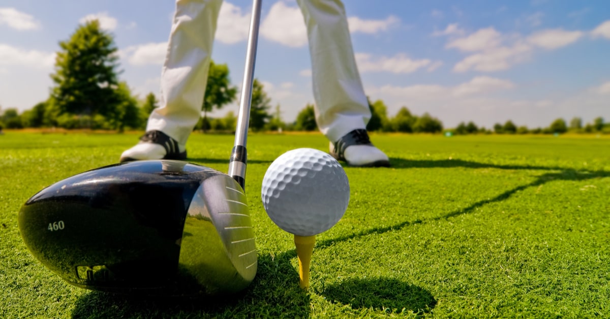 Golfball und Golfschläger auf Wiese, dahinter Beine eines Golfspielers