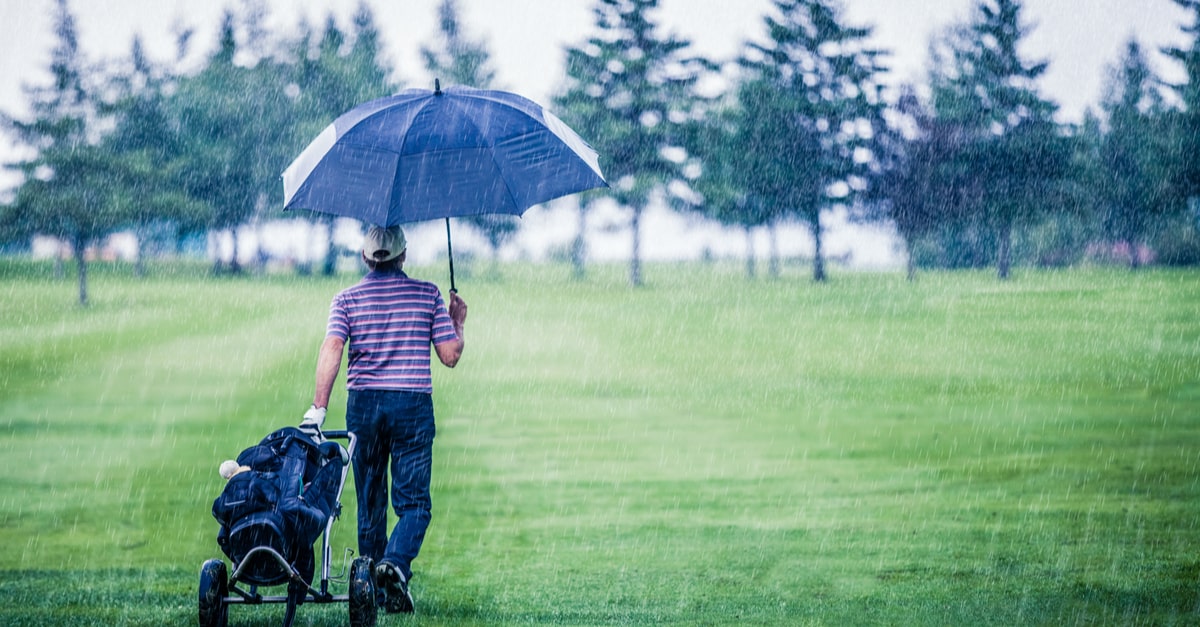 Golfer verlässt bei strömendem Regen den Golfplatz mit Golftrolley und Golfschirm